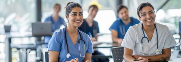 Two smiling nursing students sit at the front of their classroom listening intently and poised to take notes. Several more students, out of focus, sit behind them.