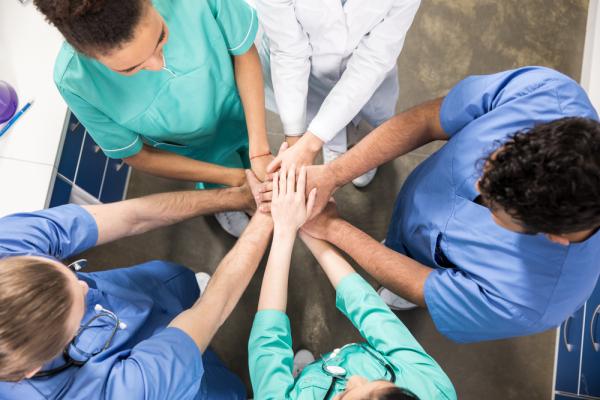 Photo of a variety of healthcare workers in their scrubs standing in a circle joining their hands in the centre of the group.