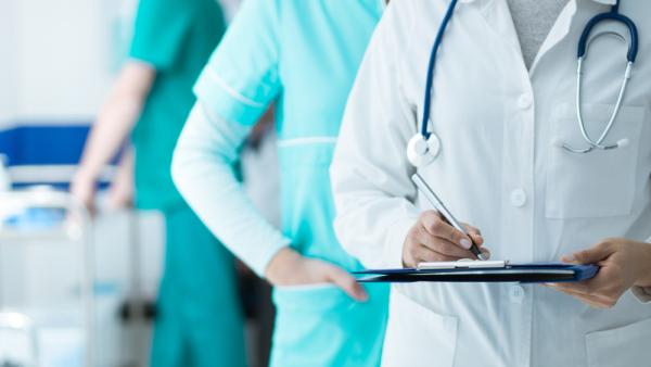Photo of people in nurses scrubs, lead by a person in a doctor’s uniform holding a pen and folder