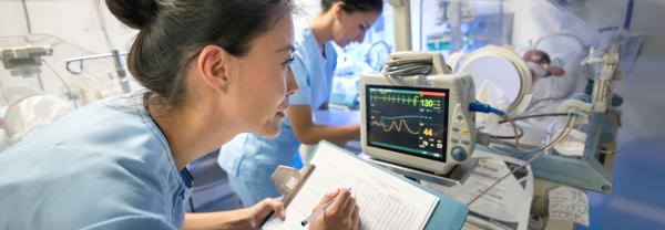 A new graduate nurse records vital signs from a monitor in the foreground, while a colleague works in the background near the patient's bed.