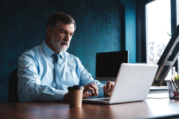 A man with a grey beard in a business shirt and tie explores online university teaching jobs on his laptop
