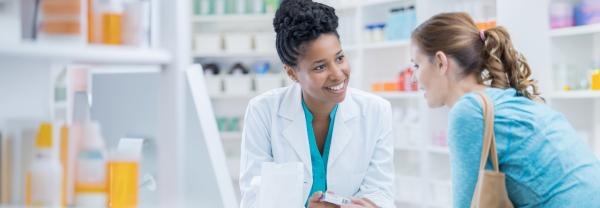 A smiling pharmacist leans against the counter as she discusses medication with her customer.