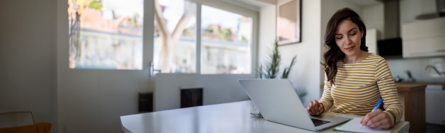 A doctoral candidate in an online program jots down notes with a pen and paper while participating in online coursework on a laptop at her kitchen table.