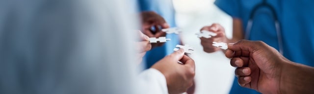 A group of unrecognizable medical professionals hold puzzle pieces near each other as though preparing to put together the pieces.
