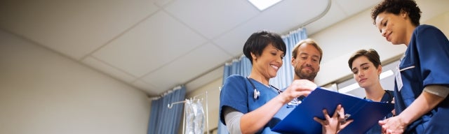 A diverse team of nurses in scrubs review documents together in a hospital room.